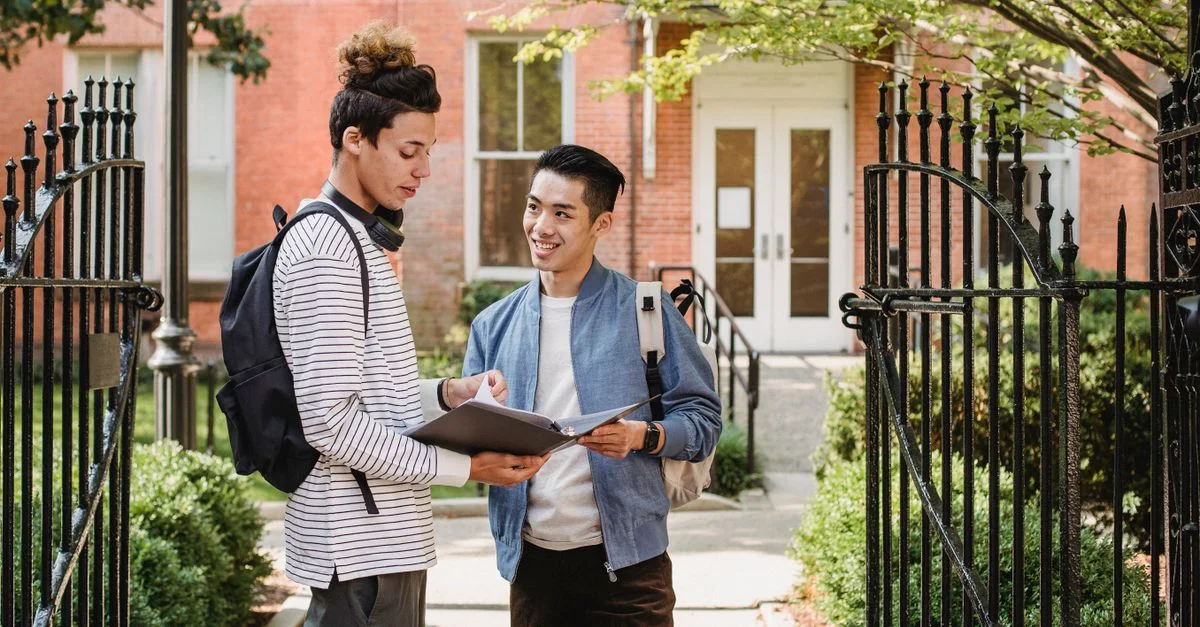 Deux étudiants discutent devant un bâtiment universitaire, partageant des notes de cours.