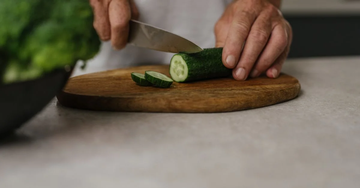 Gros plan des mains tranchant un concombre sur une planche en bois dans une cuisine.